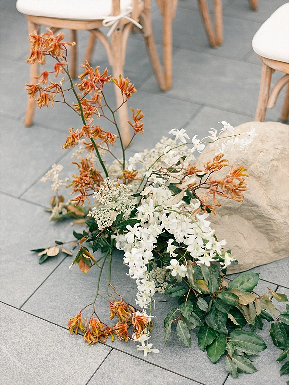 Wedding aisle florals with white and orange blooms and greenery arranged low on stone beside wooden chairs on a gray tile floor