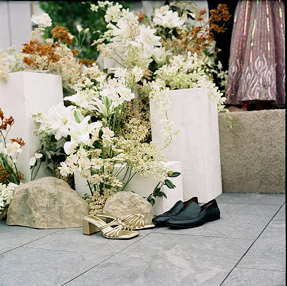 Wedding shoes with bride and groom shoes—gold heeled sandals and black loafers styled beside white florals on stone patio steps