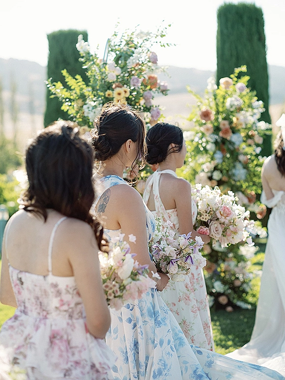 Bridesmaids processional with bridesmaids walking down aisle in floral print dresses holding pastel bouquets on a garden lawn, cypress trees behind