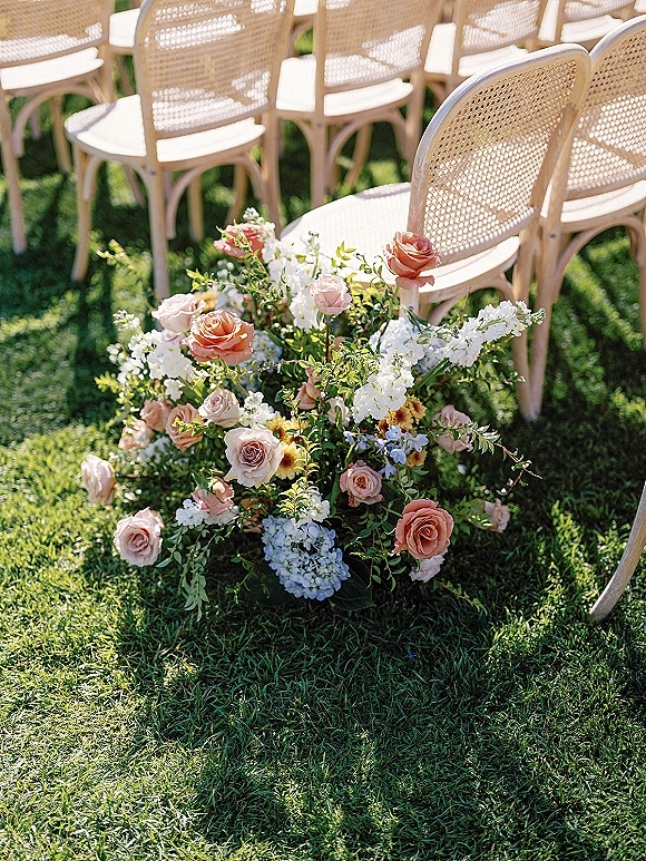 Ceremony aisle flowers in a ground floral arrangement of blush roses, white hydrangea and greenery beside rattan chairs on a grass lawn