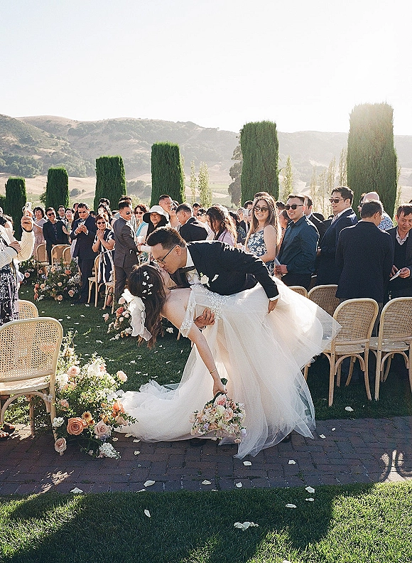 Wedding kiss moment as the couple shares a dip kiss wedding photo on a rose-petal aisle, guests cheering on a hillside lawn ceremony