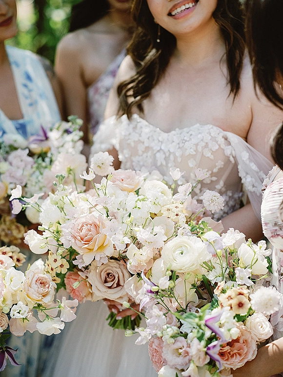 Bridesmaid bouquets of pastel flowers with roses and ranunculus, held beside a bride in lace off-shoulder gown against sunlit greenery