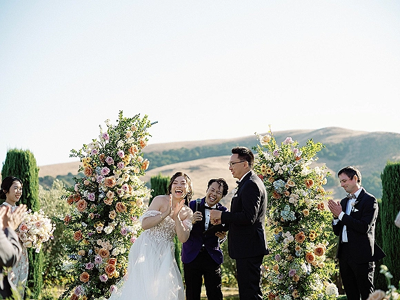 Ceremony moment as the bride laughs beside the groom under a rose and greenery arch, officiant with microphone in hillside garden setting
