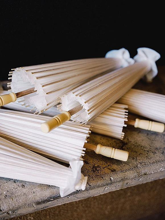 Wedding parasols and paper parasols for wedding arranged with white paper canopies, wooden handles, and ribbon ties on a stone bench by a dark wall