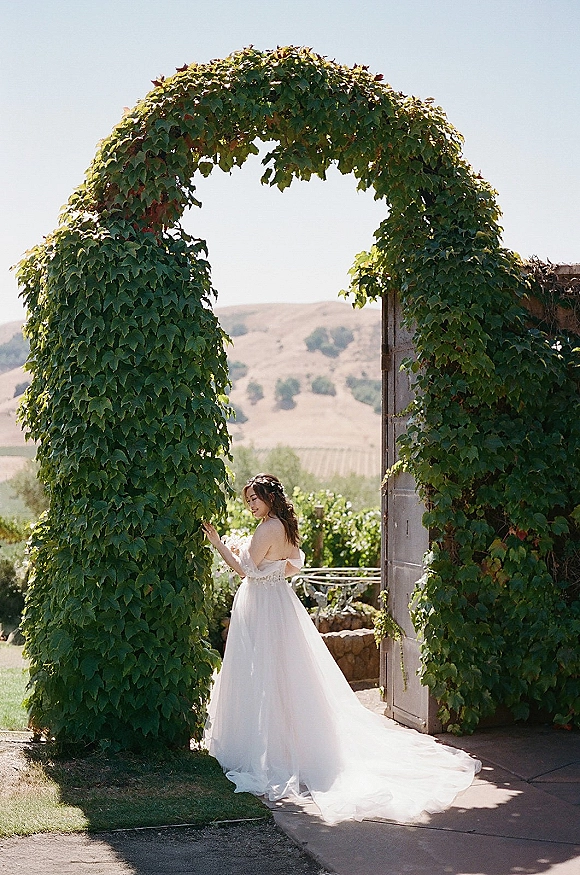 Bridal portrait of a bride in an off the shoulder gown with lace bodice and long train, looking over her shoulder under an ivy archway