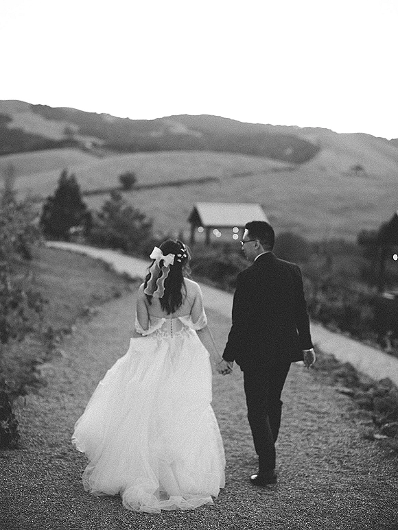Newlywed couple walking hand in hand, bride in a backless dress with a veil bow and train, groom in dark suit on a gravel path by hills and barn