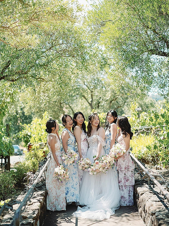 Bridesmaid group photo of the bride with bridesmaids in pastel floral dresses holding blush bouquets on a sunny garden path by a stone bridge