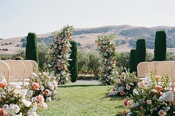 Ceremony aisle decor with pastel roses and white floral pillars lining cane-back chairs on a grass lawn with cypress trees and hills beyond