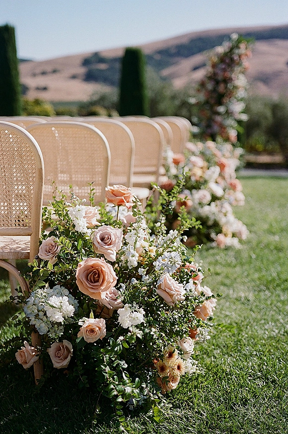 Ceremony aisle decor with wedding aisle flowers, blush and white meadow-style ground florals and cane-back chairs on a lawn with cypress trees