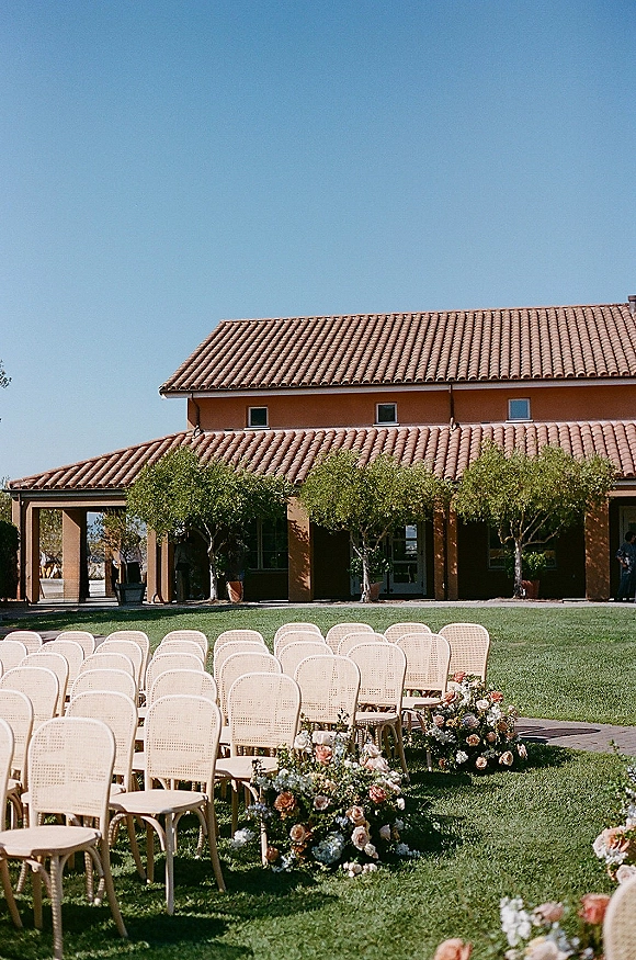 Ceremony setup with rattan wedding chairs in rows, lined by aisle florals on a grass lawn beside a terracotta-roof villa veranda under blue sky