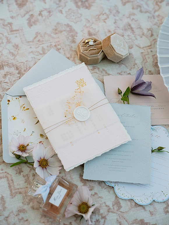 Wedding invitation suite with wax seal, deckled edge cards, floral illustrations, rings, and ribbon arranged on patterned linen fabric