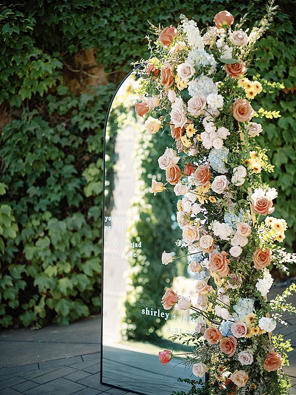 Wedding welcome sign on an arched mirror with pastel rose and hydrangea flowers, set by an ivy wall along a stone garden walkway