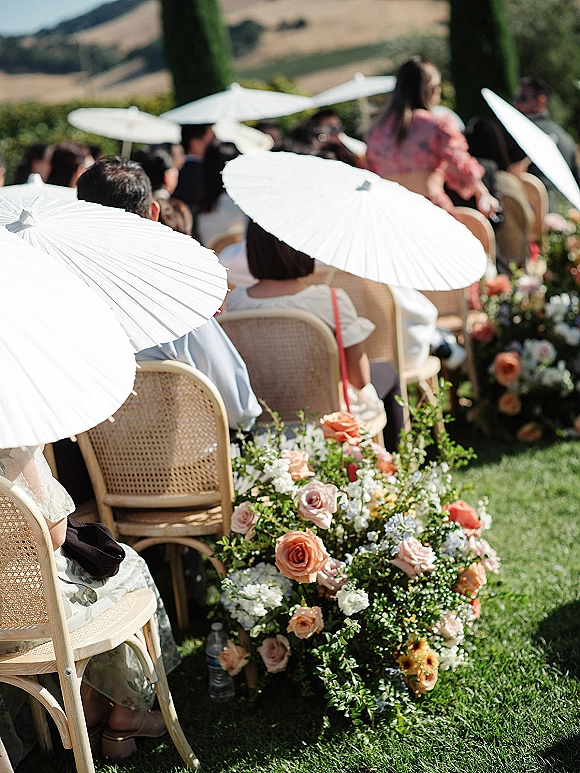 Ceremony guest seating with outdoor wedding ceremony chairs, cane bistro rows and white paper parasols along rose-and-greenery aisle on lawn hills