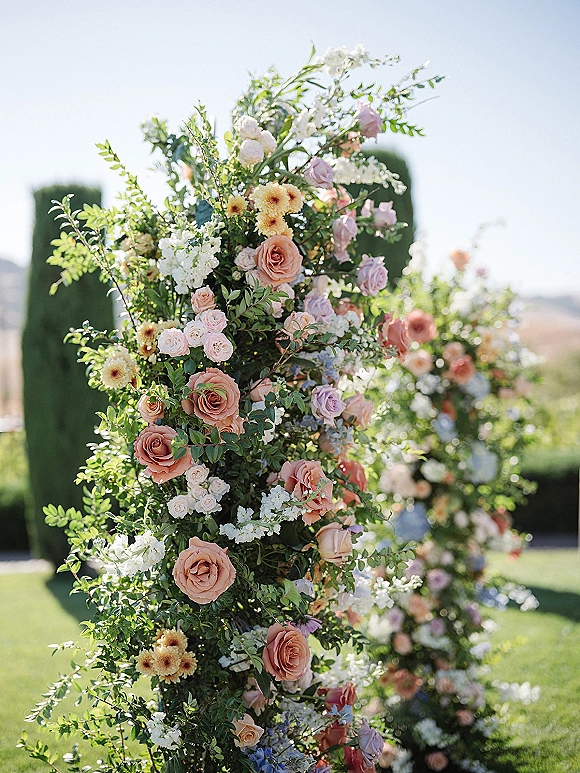 Wedding floral arch filled with roses and garden flowers, draped in greenery on a lawn with trimmed hedges under open sky