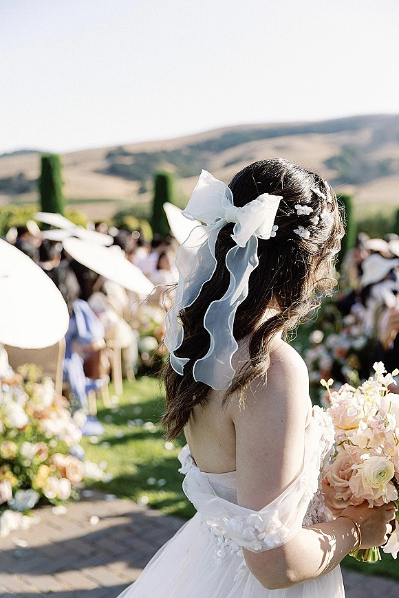 Bridal portrait of a bride from behind with a white hair bow, holding a bouquet on an outdoor ceremony aisle with guests under umbrellas