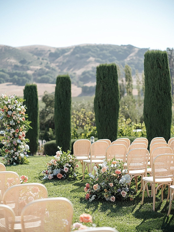 Outdoor ceremony setup with rattan chairs and garden wedding ceremony aisle lined with roses and hydrangeas on grass, cypress trees beyond