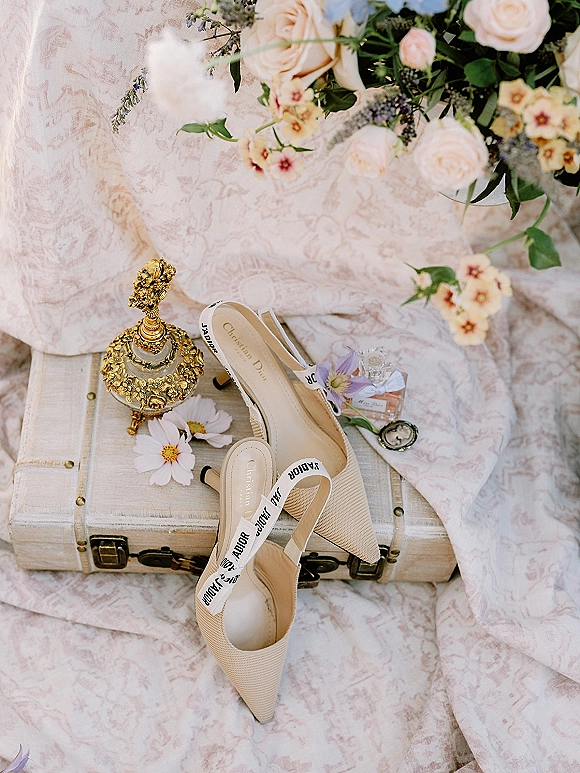 Bridal shoes flatlay with wedding shoe flatlay slingback heels, vintage perfume bottle, bouquet and cameo pendant on patterned fabric backdrop