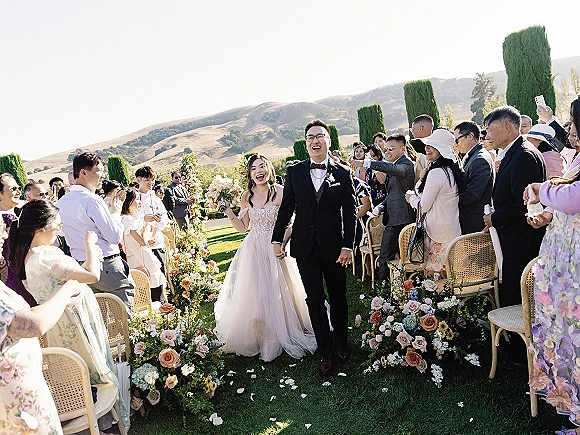 Wedding recessional as bride and groom walk hand in hand down the aisle, bouquet in her strapless lace dress, guests cheering on a sunny lawn