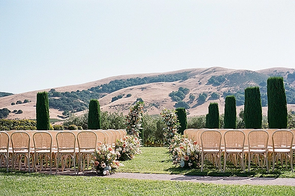 Ceremony setup for an outdoor wedding ceremony with asymmetrical floral arch and pastel aisle markers on a lawn with hills and cypress trees
