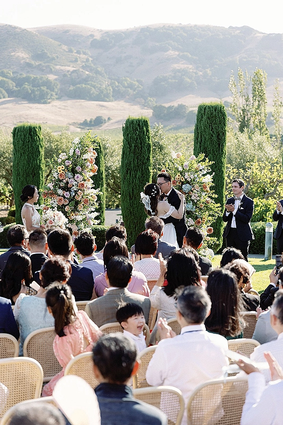 Ceremony kiss as the bride and groom kiss beneath a rose-and-greenery floral arch, guests clapping on a sunny lawn with mountains behind