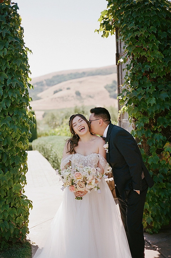 Couple portrait of groom kissing bride’s cheek as she laughs, holding a pastel bouquet under a vine-covered archway on a garden walkway