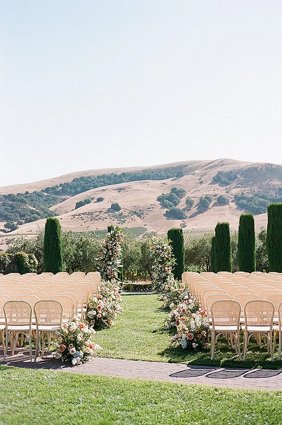 Ceremony setup with pastel wedding florals lining the aisle and floral pillars, cane-back chairs on a lawn amid vineyard rows and hills.