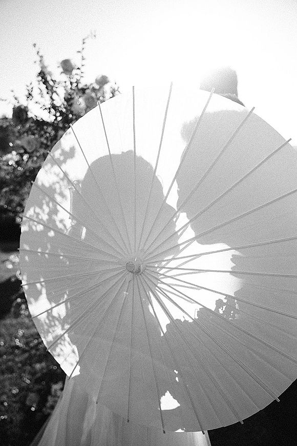 Wedding couple portrait with paper parasol, bride’s veil and groom’s suit jacket silhouetted against sunlit trees and bright sky