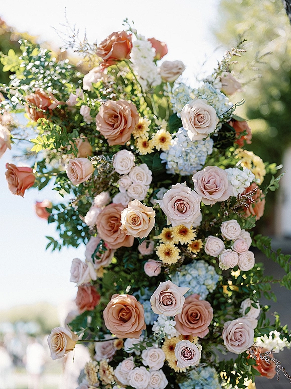 Wedding floral arrangement on a ceremony floral arch with blush roses and hydrangea, lush greenery, and soft garden sunlight in back