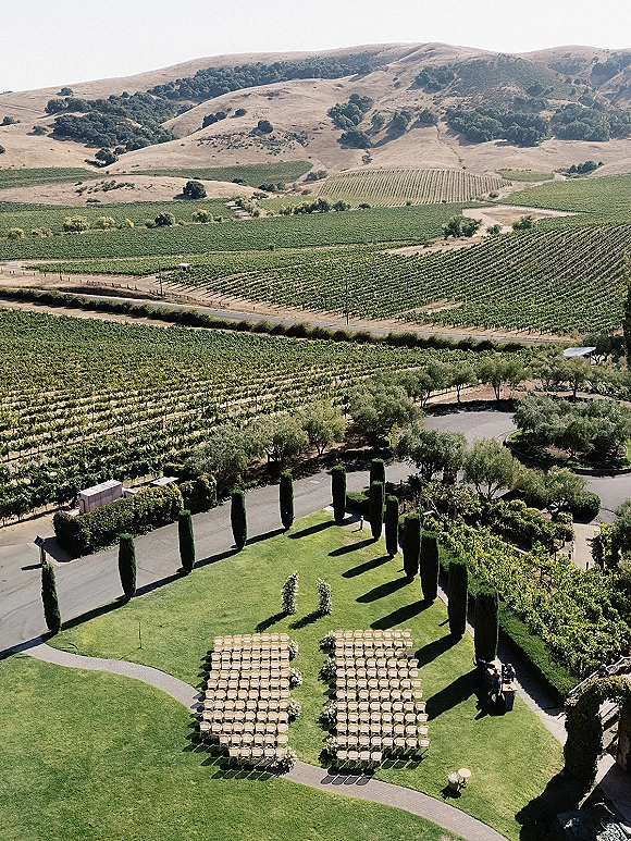 Outdoor ceremony setup with vineyard wedding ceremony chairs along a curved aisle, floral clusters and greenery hedges overlooking vineyard rows and hills