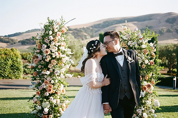 Wedding couple portrait of bride and groom kissing beneath a floral arch, her veil and off-shoulder dress against lawn and hills backdrop