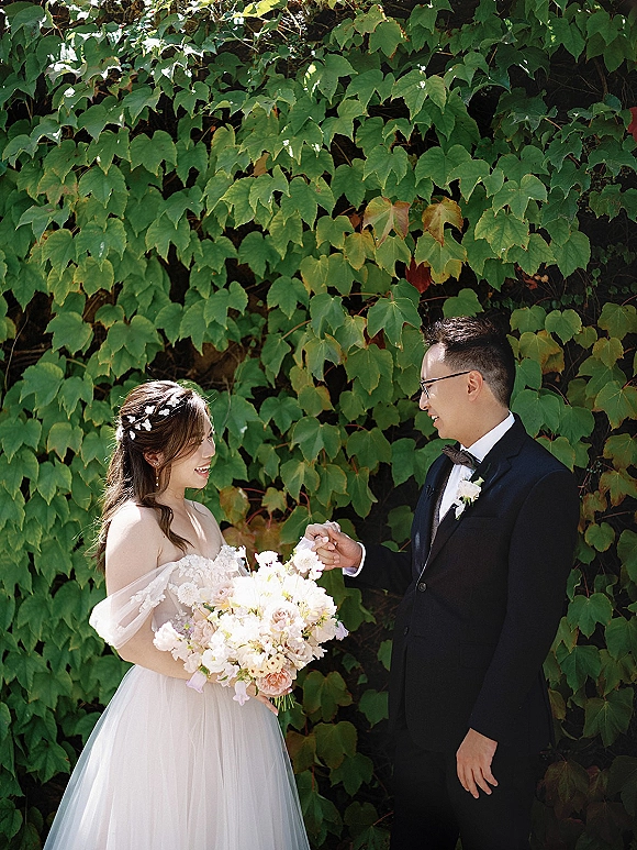 Couple portrait of bride and groom laughing as she holds a pastel bouquet, his black tuxedo and boutonniere against an ivy wall backdrop