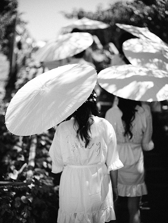 Bridesmaids getting ready with bridal party parasols, wearing matching embroidered robes and waist ties in a sunlit garden under trees