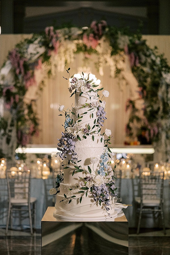 Wedding cake with tiered wedding cake design, cascading sugar flowers and greenery vines on a pedestal beside candles under string lights
