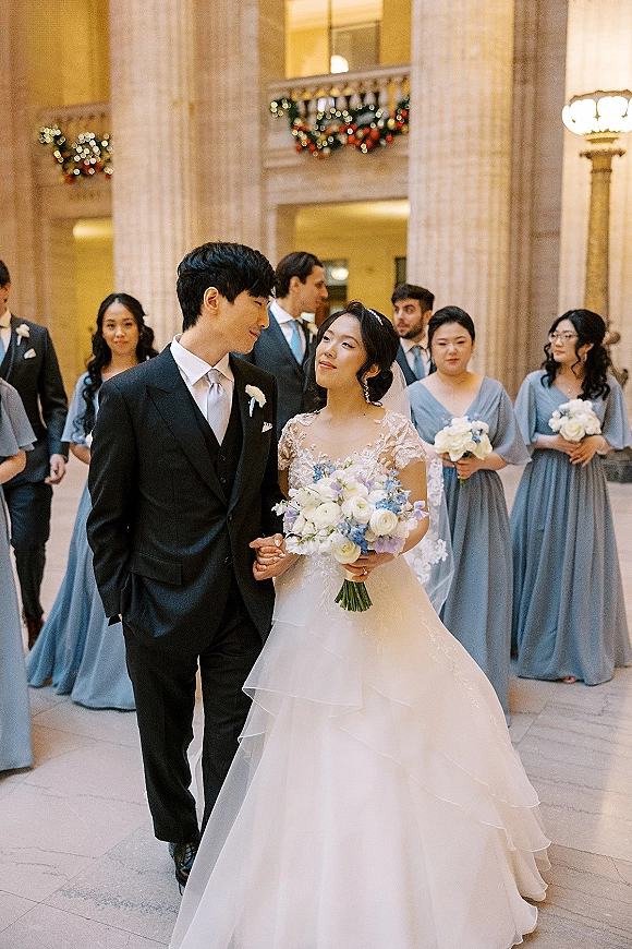 Wedding party portrait with bride holding bouquet beside groom in suit, bridesmaids in blue dresses in a grand columned hall