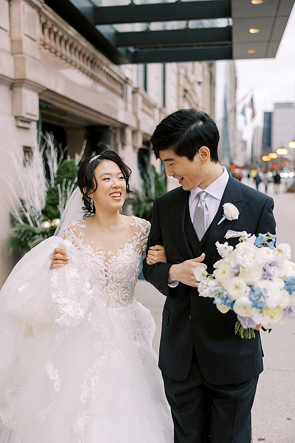 Couple portrait of bride and groom walking and laughing on a city sidewalk, bride in veil holding her gown beside him under street lights