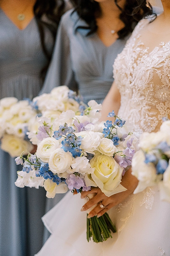 Bridal bouquet of white roses and blue delphinium with ranunculus and lavender, held beside a lace wedding dress in soft indoor light