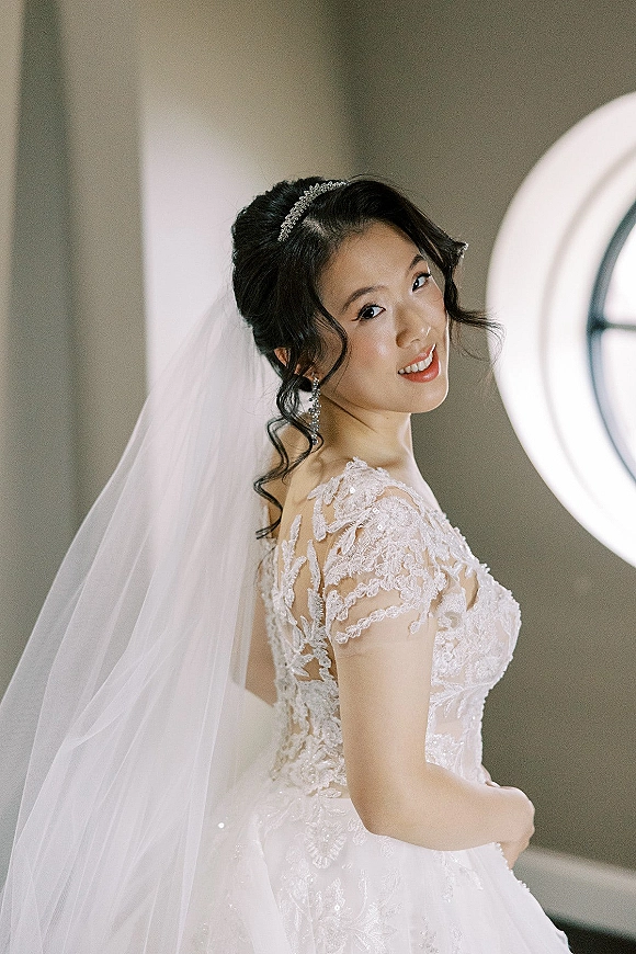 Bridal portrait of a bride looking back in a lace wedding dress with cathedral veil and tiara headband by an arched window in natural light