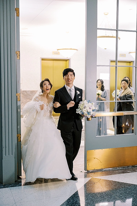 Wedding recessional as bride and groom walk arm in arm through a hotel lobby hallway, bride holding gown, veil trailing past guests