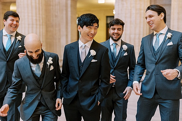 Groomsmen portrait of the groom with groomsmen laughing in suits and vests with boutonnieres in a warm-lit hallway with columns