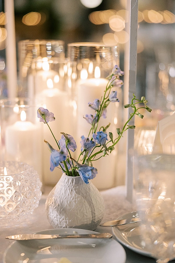 Reception tablescape with wedding table centerpiece of blue flowers in a white bud vase, pillar candles, and place settings under string lights