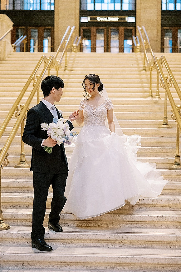 Couple portrait of bride and groom on stairs, holding hands by a grand staircase with brass railings; bride in veil with bouquet
