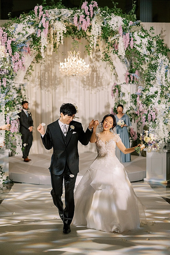 Wedding recessional as bride and groom walk the aisle under a floral arch with chandelier, petals falling, and bridesmaid in dusty blue behind