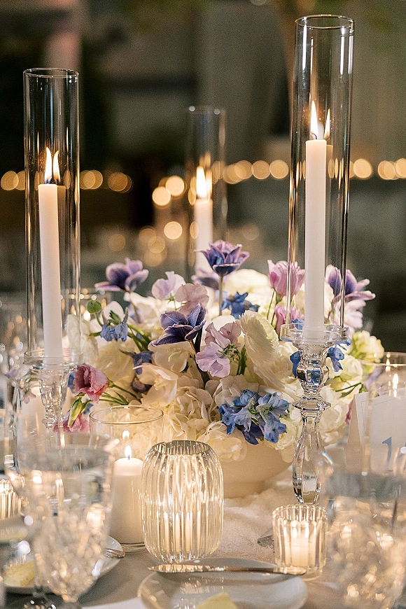 Wedding tablescape with candlelit reception table featuring tall glass candle holders, taper candles, low floral centerpiece, and bokeh lights