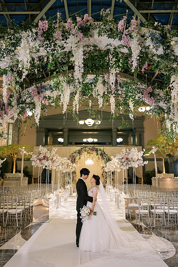 Wedding couple portrait of bride and groom embrace on a candle-lined white aisle runner beneath hanging florals and a chandelier in a grand hall