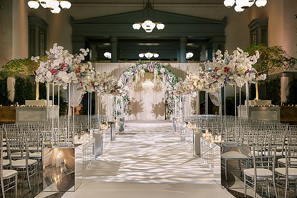 Ceremony setup with indoor wedding ceremony setup featuring a candle-lined aisle runner, floral arch, drapery, and chandeliers in a columned hall
