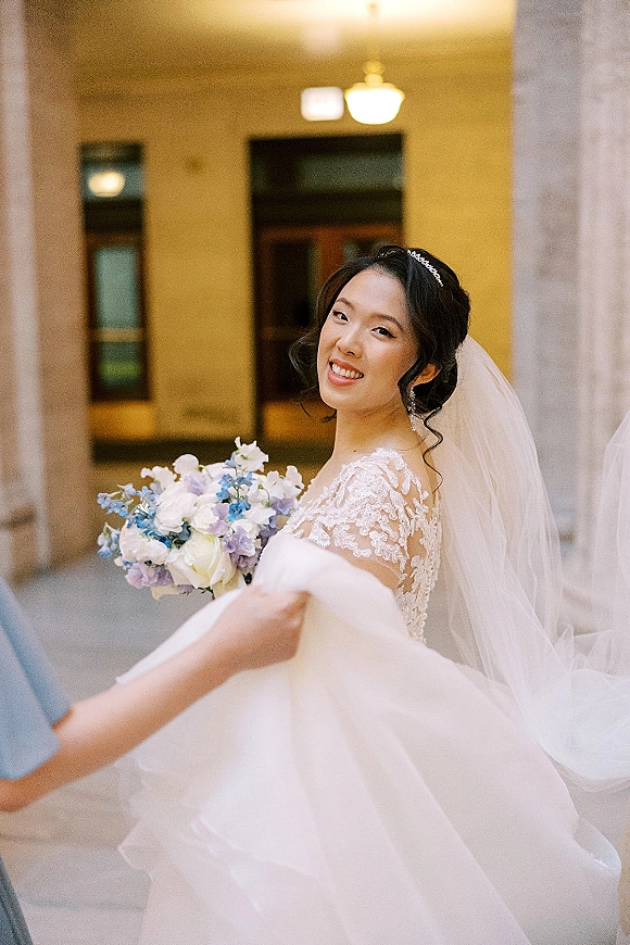 Bridal portrait of a bride holding bouquet, looking back with veil over shoulder in a stone-column hallway under a chandelier