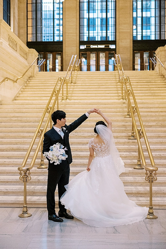 Couple portrait of bride twirling in lace gown and veil as groom watches, holding a white and blue bouquet on a grand staircase
