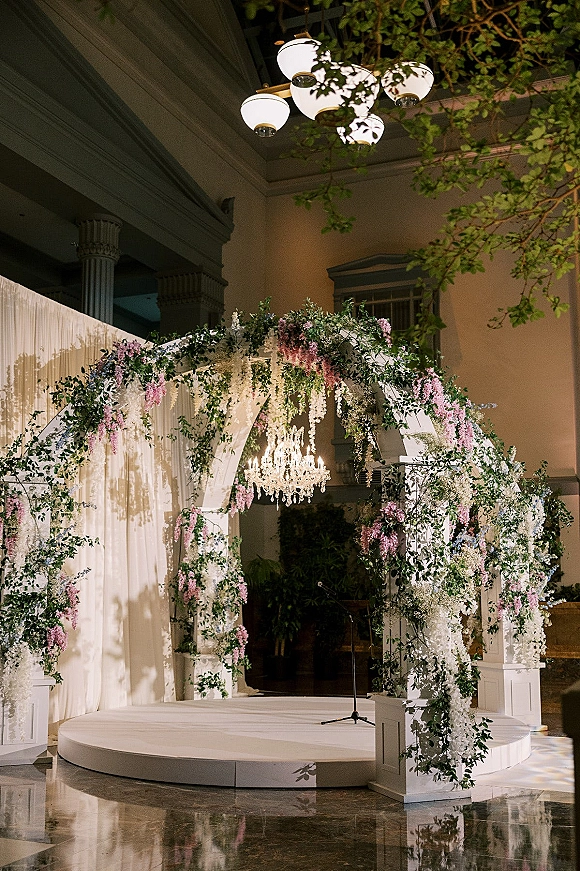 Wedding altar decor with a floral wedding arch of hanging wisteria and greenery, white drapery and chandelier on a round stage in a grand hall