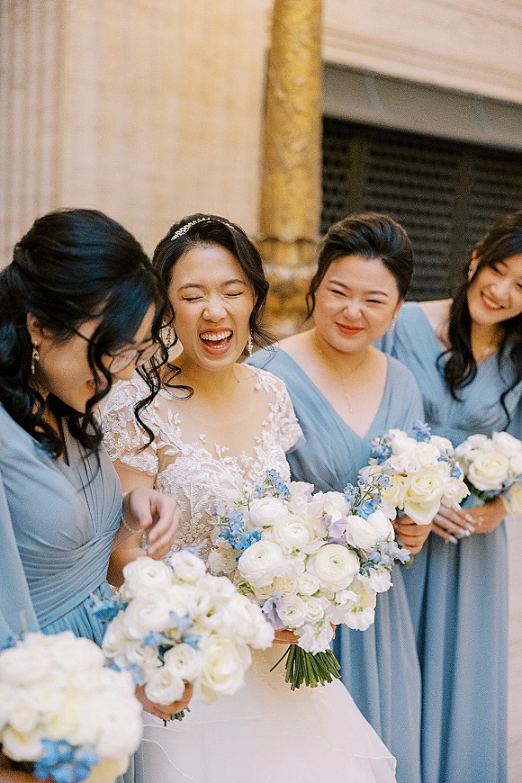 Bride with bridesmaids laughing together, holding white rose and blue bouquets by stone columns and a dark doorway outside a building
