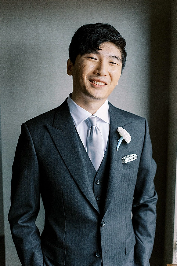 Groom portrait in a three piece suit, wearing a necktie and white rose boutonniere, lit by window light against a neutral wall